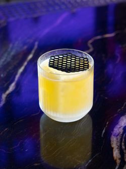 Yellow cocktail served in a textured clear glass with a black decorative garnish on top, placed on a dark purple marbled bar.