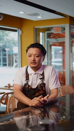 Man wearing a Jazba apron sitting at a bar counter inside an East Village Indian restaurant with modern decor.