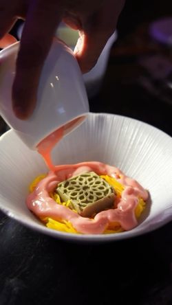 A hand pours pink sauce over a dish with yellow noodles and a decorative green square garnish in a white bowl.
