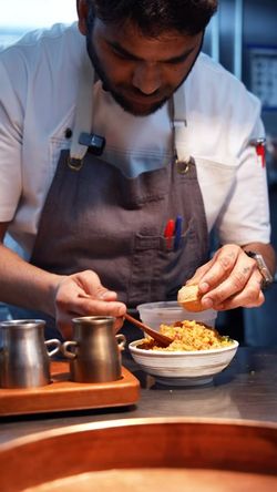 Chef in apron preparing traditional Indian dish with rice, adding garnish in restaurant kitchen setting.
