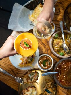 Top view of Indian food dishes on a wooden table with hands holding a cocktail and a glass of water above the plates.