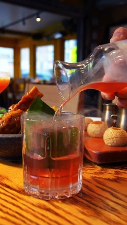 Cocktail being poured into a glass with Indian appetizers including fried snacks and pani puri on a wooden table in a dimly 