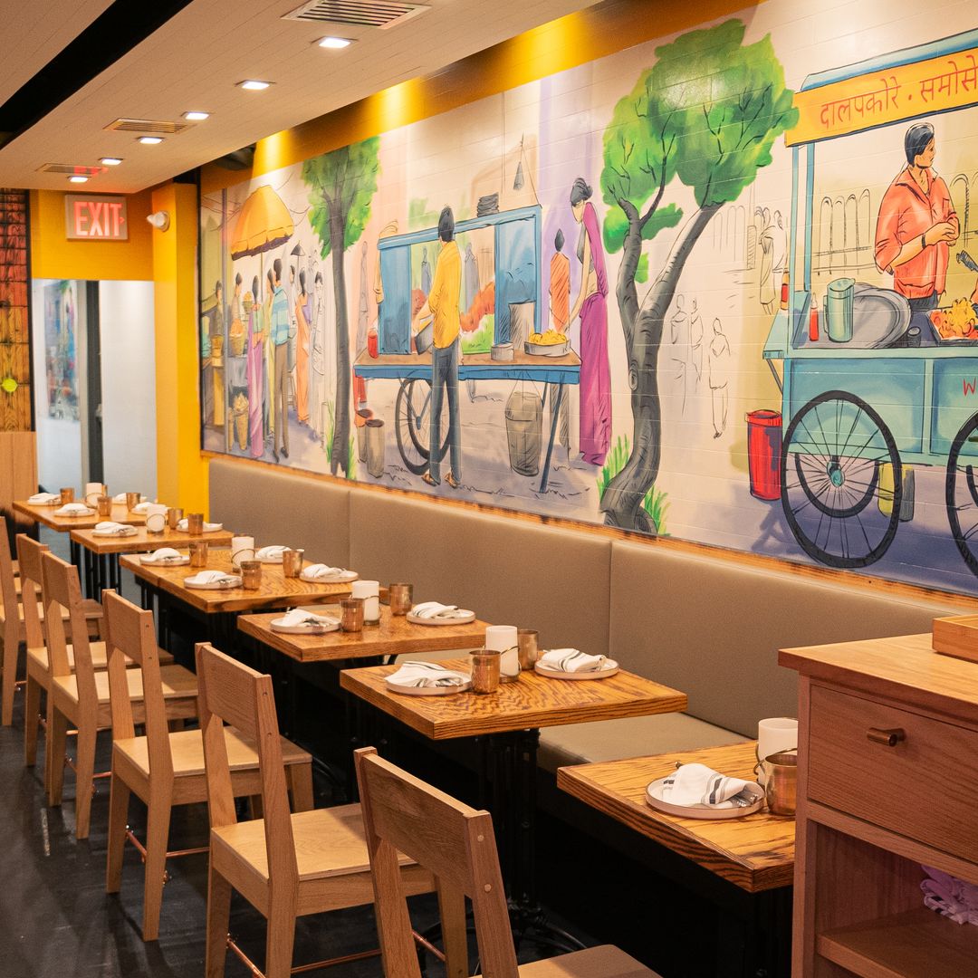 Dining area at Jazba featuring wooden tables, chairs, and a colorful mural of Indian street food vendors on the wall.
