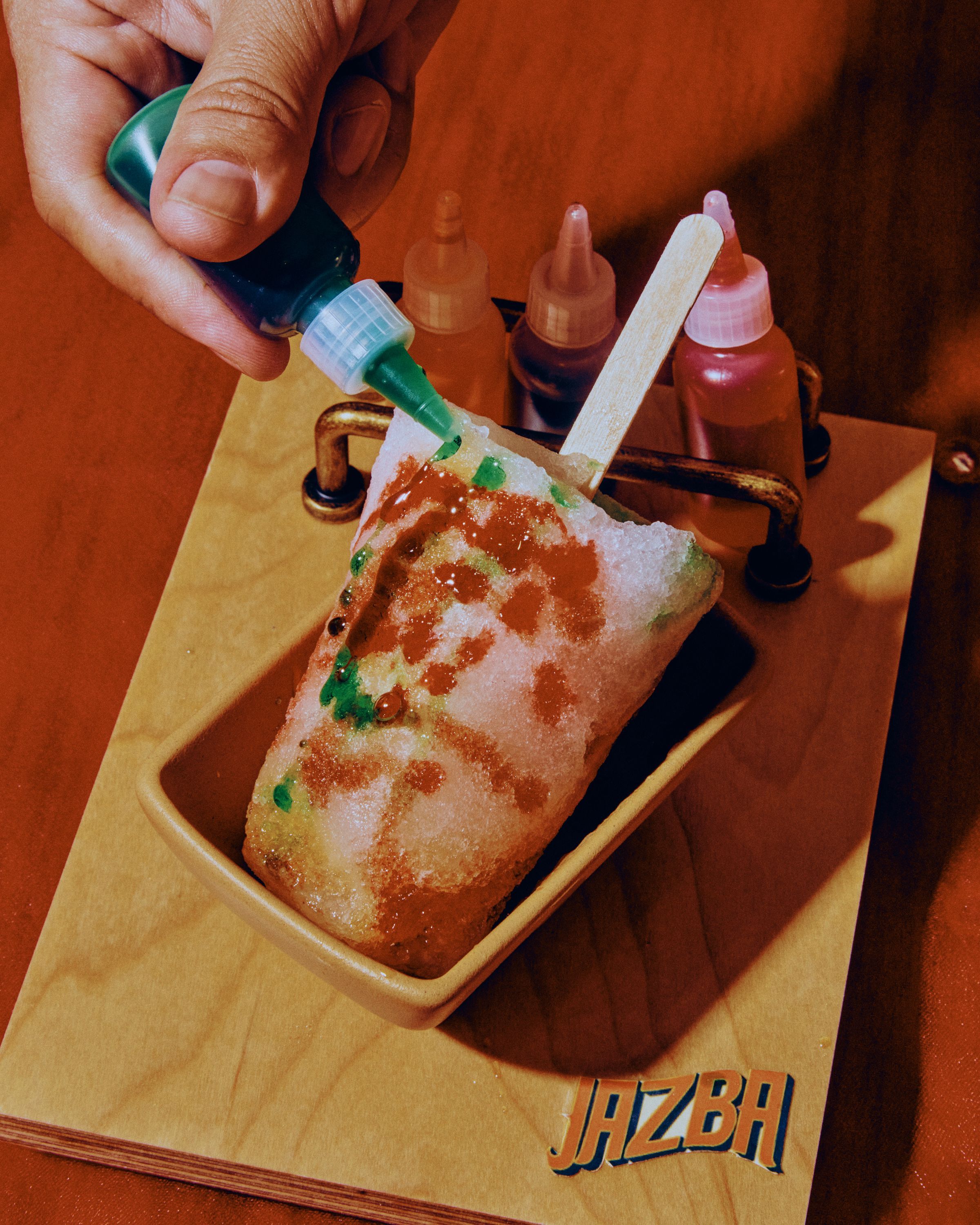 Colorful ice pop being decorated with green syrup in a container at Jazba, an Indian cuisine restaurant.