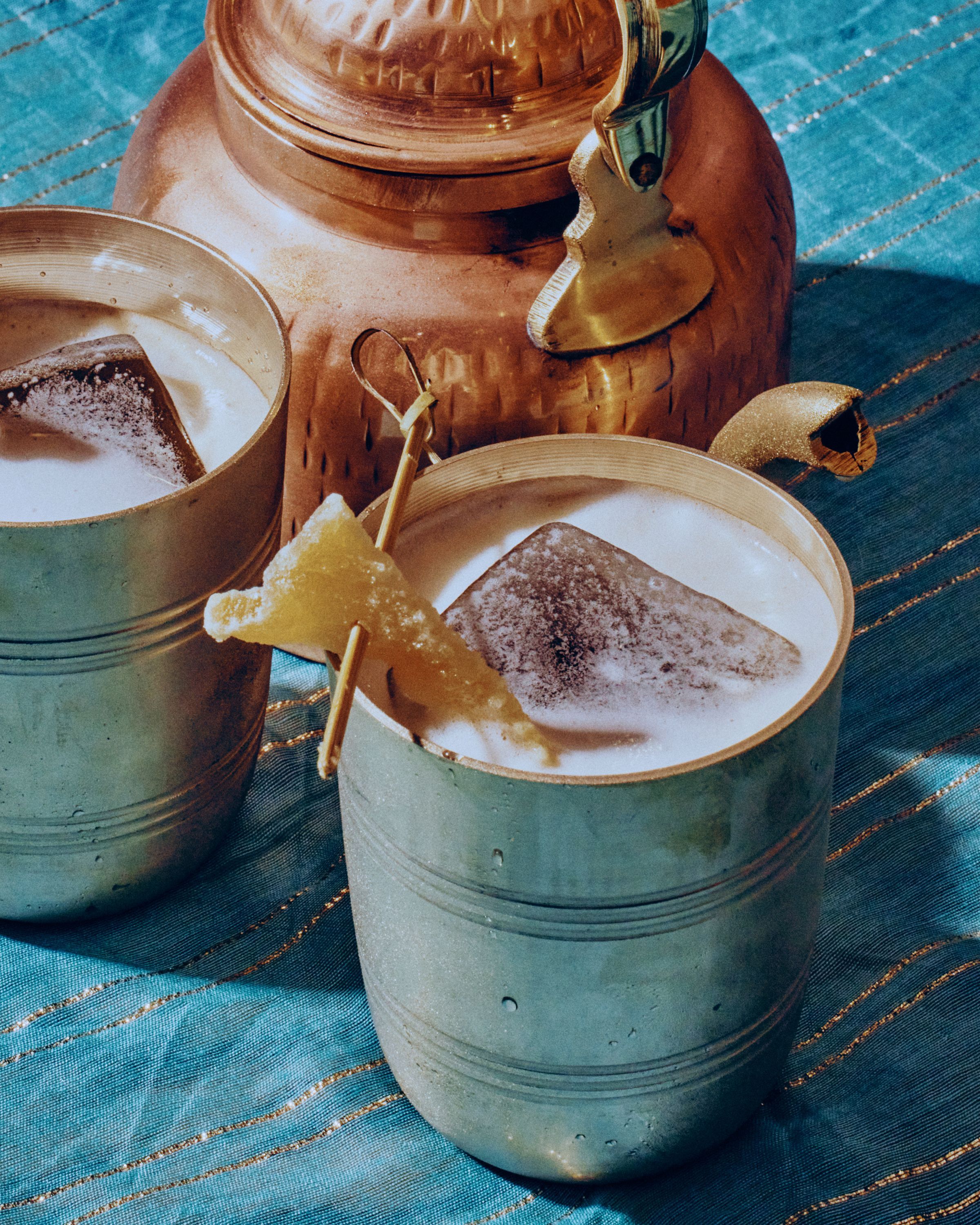 Traditional Indian drinks served in metal cups next to a copper kettle on a textured blue cloth at Jazba restaurant.