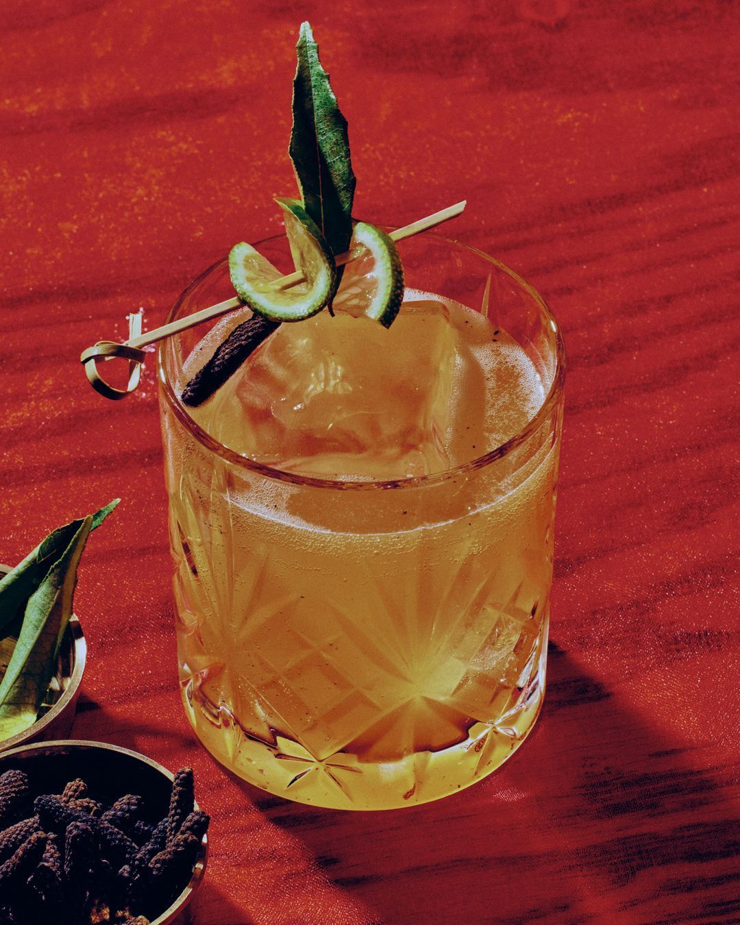 Cocktail in a glass with ice, garnished with lime slices and a green leaf on a red wooden surface.
