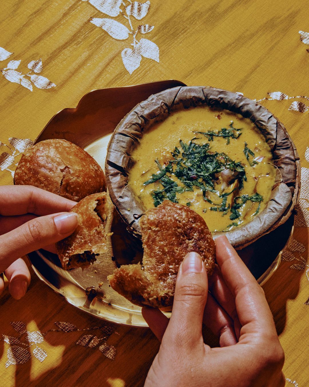 Traditional Indian dish served in a leaf bowl with hands breaking bread on a yellow patterned tablecloth at Jazba