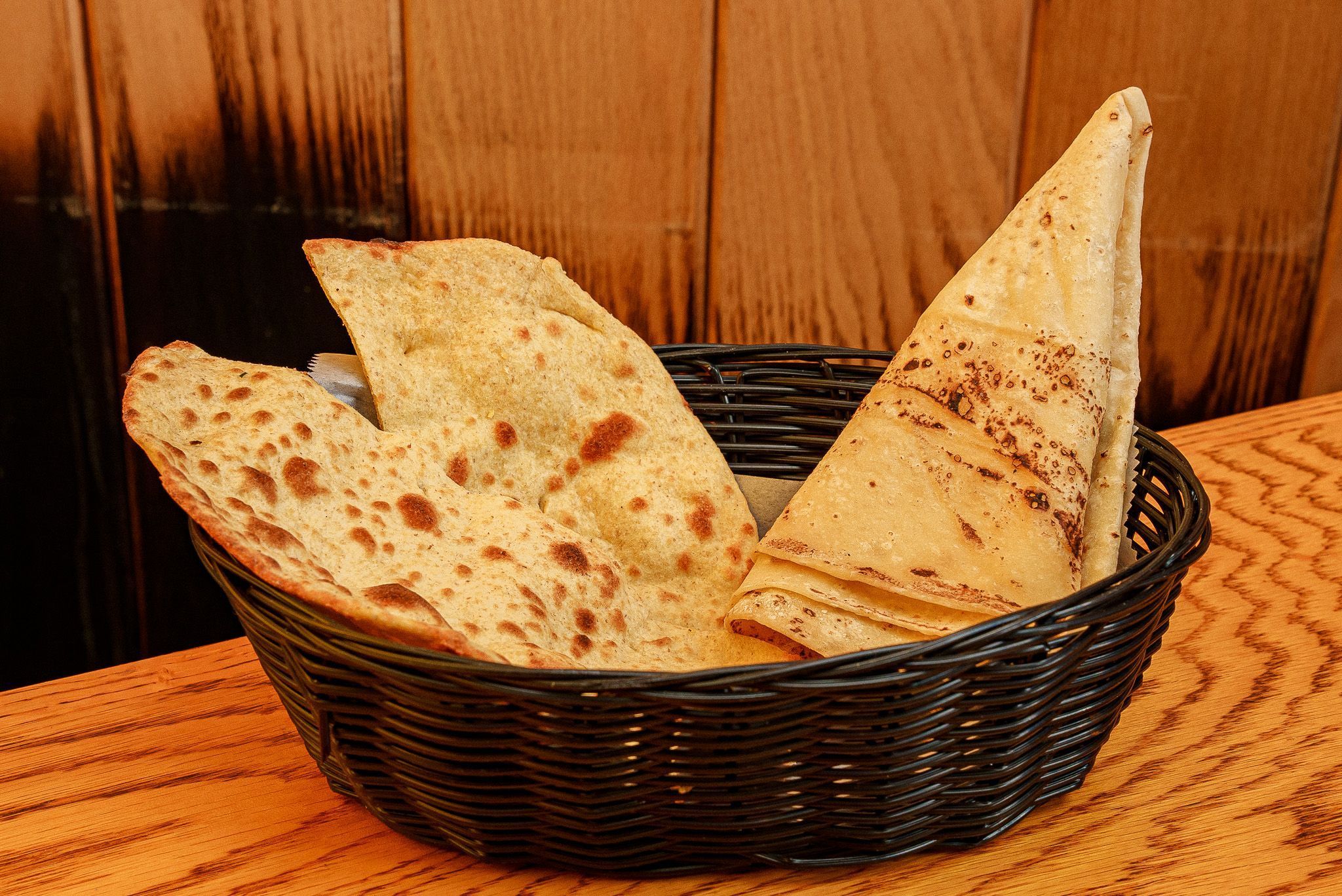 Two types of Indian flatbread, including naan and roti, served in a black basket on a wooden table at Jazba restaurant.
